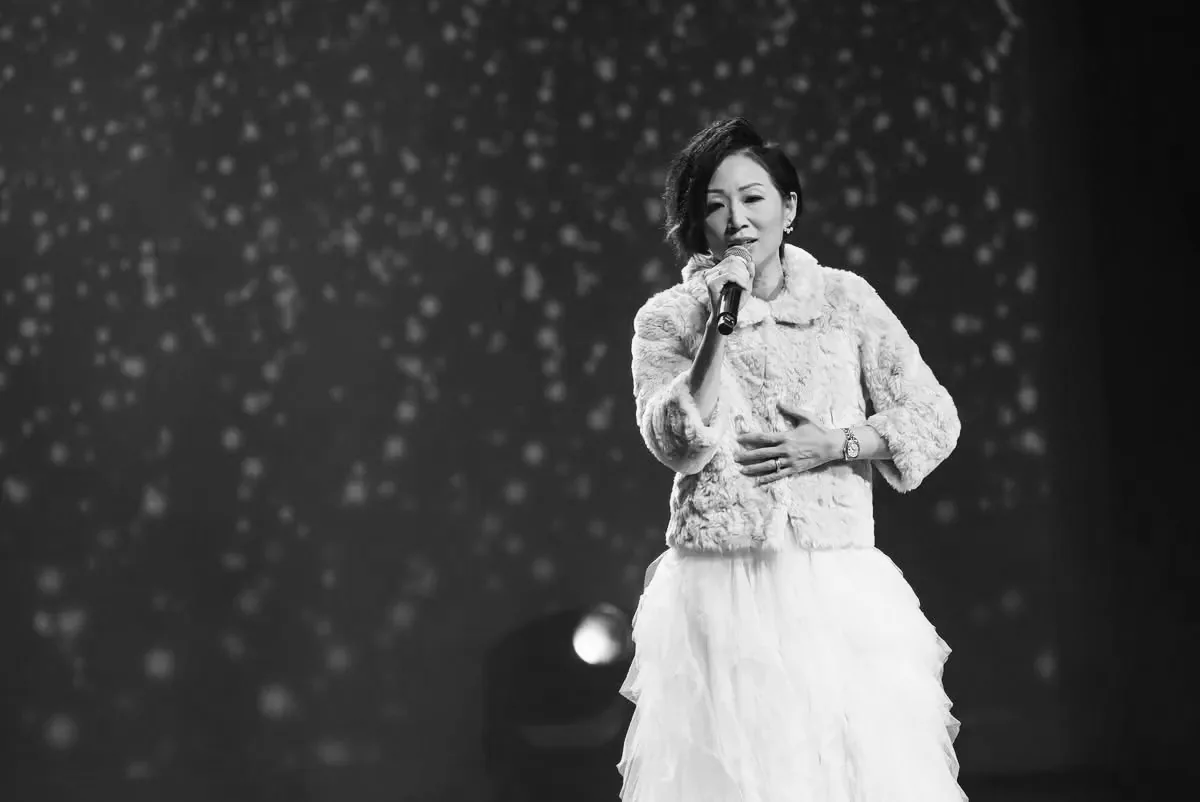 Anita Tong singing on a concert stage in a fur jacket and tulle skirt, a starlit projection behind her