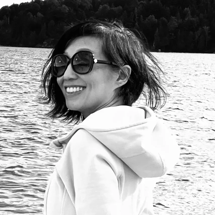 Anita Tong souriant par-dessus son épaule, lunettes de soleil, un lac et une rive boisée derrière elle