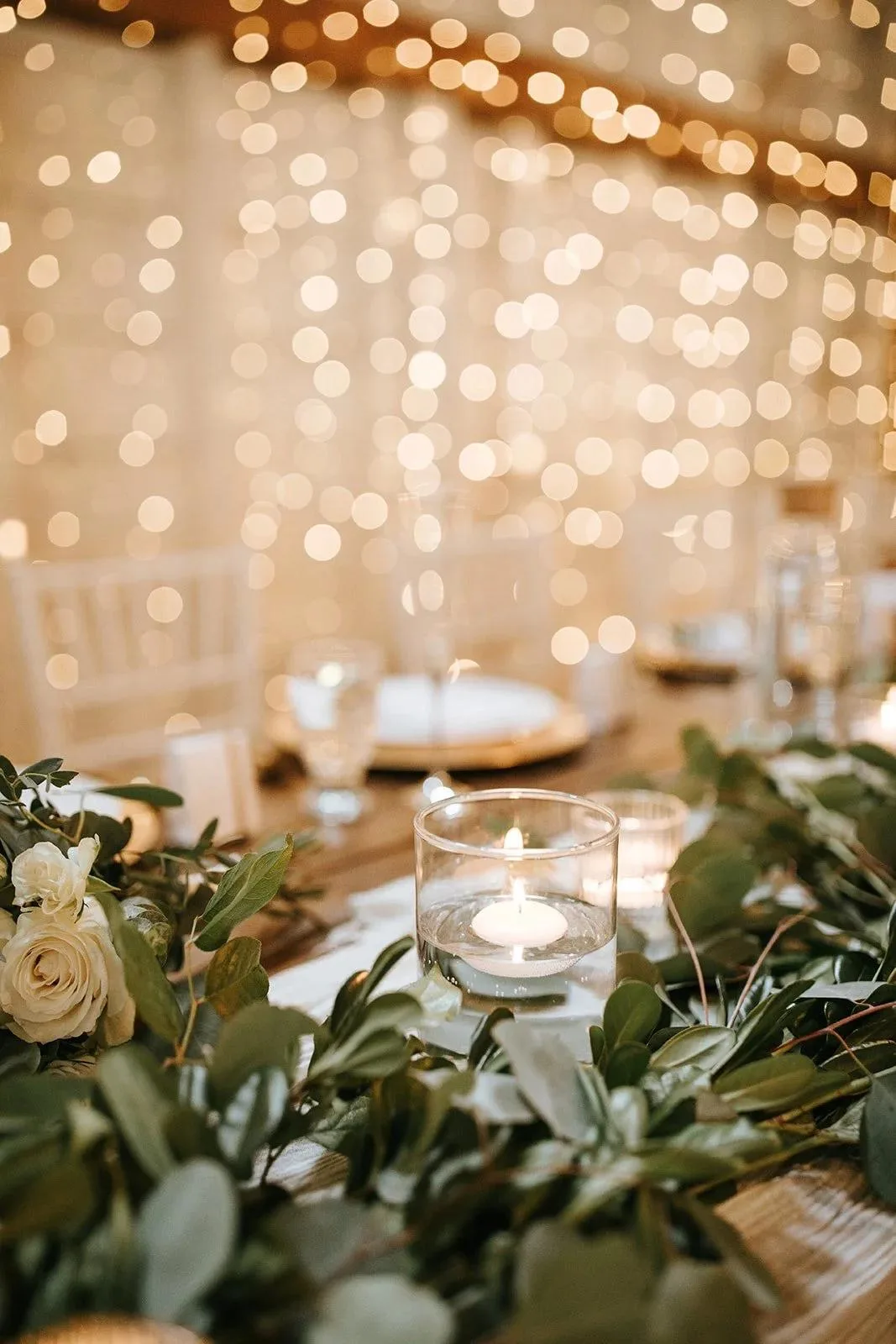 Candlelit wedding table set with white roses and greenery beneath strings of fairy lights