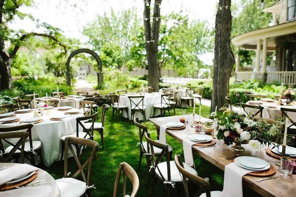 Wedding tables set on a garden lawn beneath mature trees, a stone arch beyond